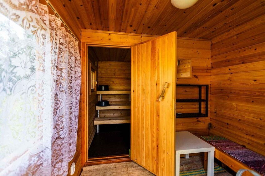 A wooden cabin interior with a bed, a table, and a window with lace curtains. 4