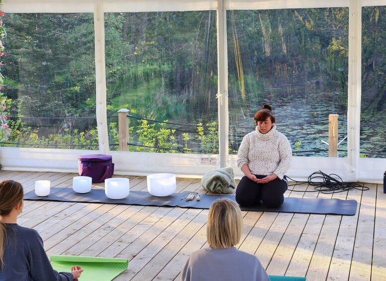 A person sitting on a yoga mat in a wooden deck area with a view of a forest and water. 2