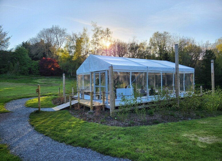 A glass greenhouse structure with a wooden deck, surrounded by a gravel path and lush greenery. 3
