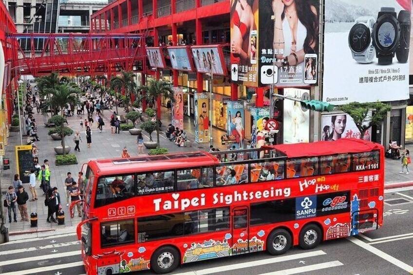 A red double-decker bus with 'Taipei Sightseeing' and 'Hop On Hop Off' advertisements, driving through a busy urban area with pedestrians and buildings. 1