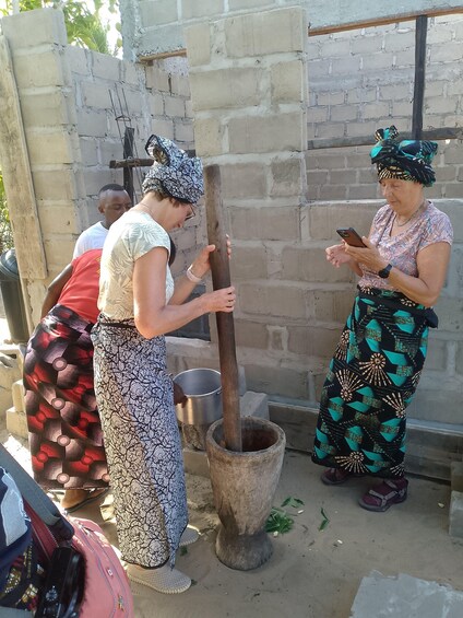 Two women grinding grain using a traditional mortar and pestle, with a metal pot nearby. 3