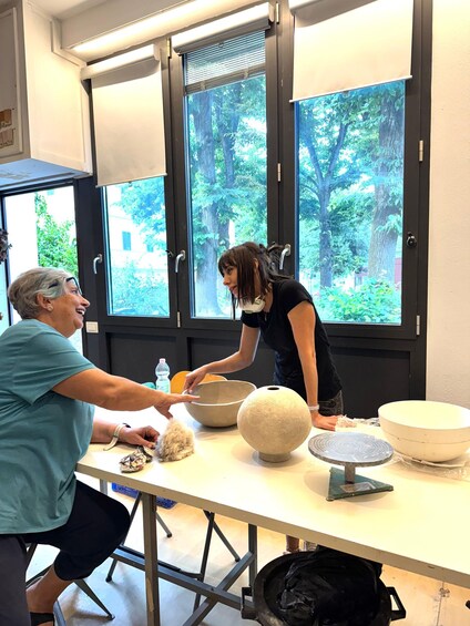 A person kneeling at a table with bowls and a scale, another person standing beside them, and a window with a view of trees. 4