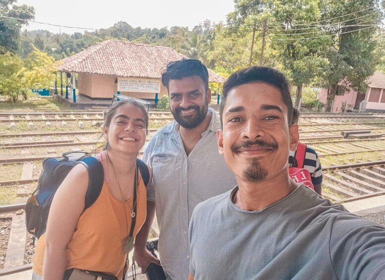 Three people standing by railway tracks with a station building in the background. 1
