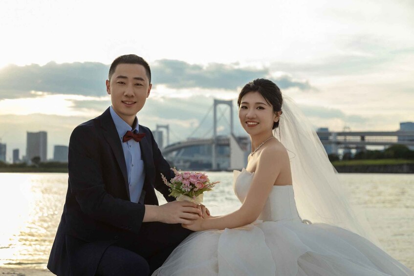 A bride and groom seated by a river with a bridge in the background. 1