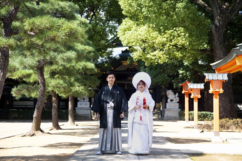 Two individuals in traditional attire standing in a park with trees and a pagoda in the background. 3