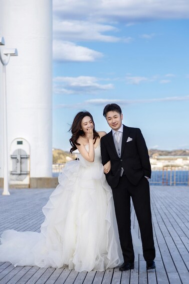 A couple in wedding attire on a wooden deck with a white building and a clear sky in the background. 2