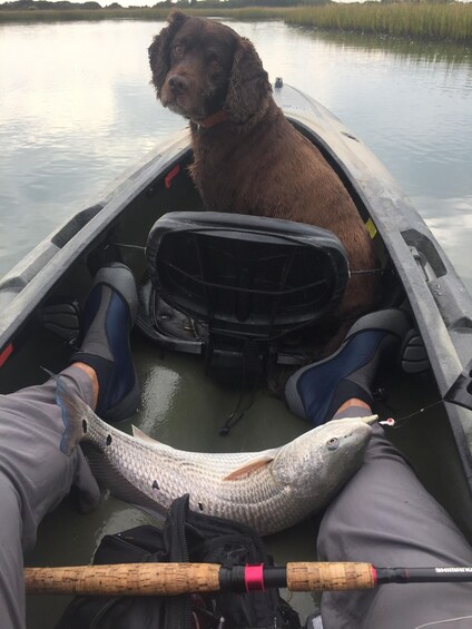 A person fishing with a large fish on a boat, a dog sitting in the boat, and a calm water body in the background. 2