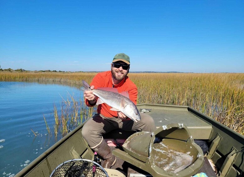 A person holding a fish in a boat on a calm water body surrounded by tall grasses. 1