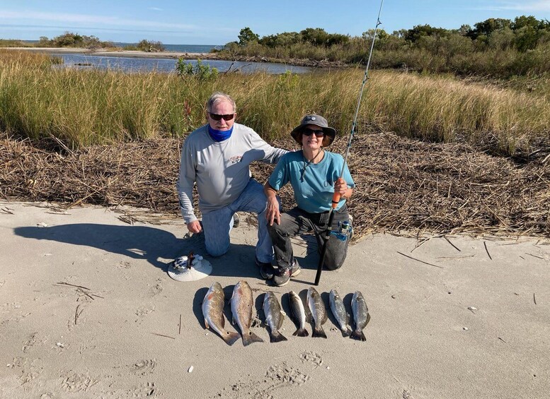 Two people fishing by a sandy beach with a body of water and grass in the background. 3