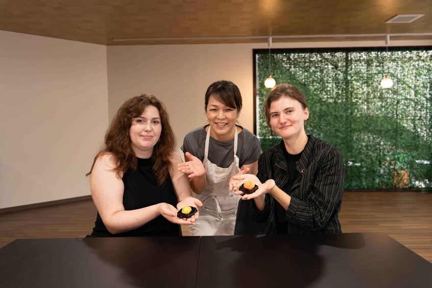 Three people holding small food items at a table in a room with a wooden floor and a green wall. 3