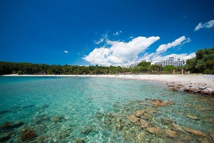 A clear turquoise sea, a sandy beach, and a forested hill with buildings in the background. 2