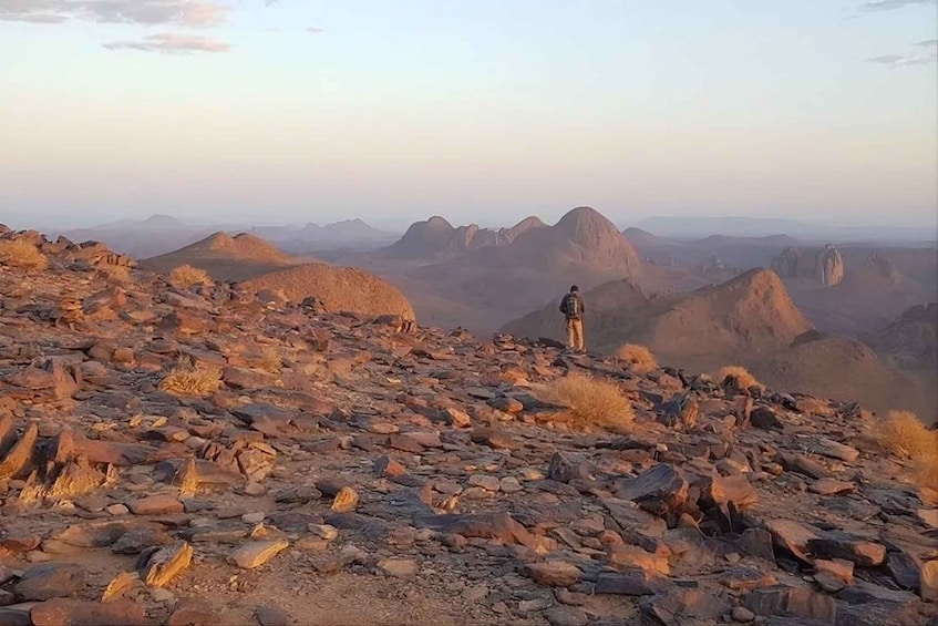 A rocky terrain with a person standing on a hill, overlooking a range of mountains. 1
