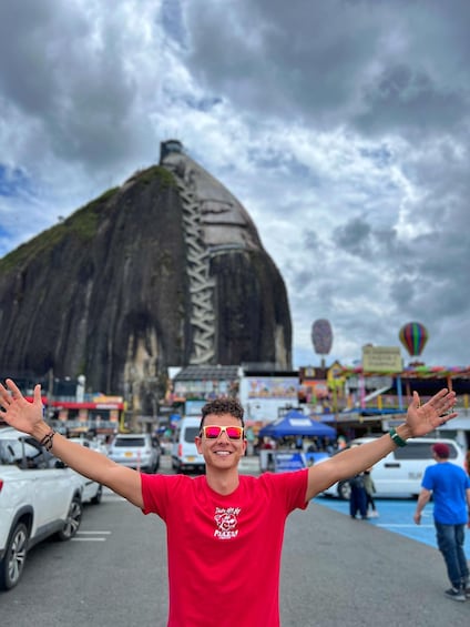 A person in a red shirt with arms outstretched, standing in front of a large rock formation with a cloudy sky in the background. 1