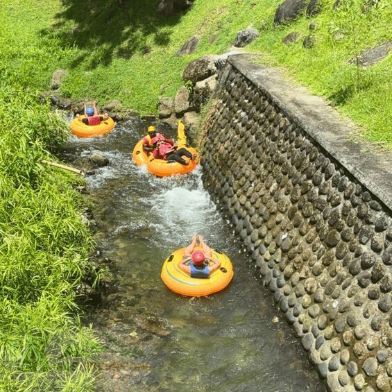 People floating on inflatable tubes in a narrow water channel surrounded by greenery and a stone wall. 1