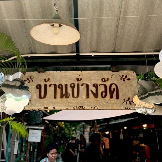 Een bord met Thaise letters, hangend in een markt met hangende lampen en planten. 3