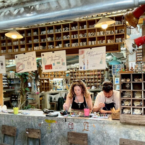 Twee personen schilderen aan een toonbank, omringd door diverse kunstbenodigdheden en een plank met lettermagneetjes op de achtergrond. 1