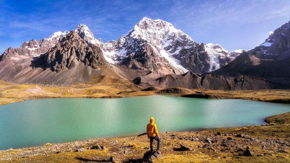 A person in a yellow jacket standing by a turquoise lake with snow-capped mountains in the background. 5