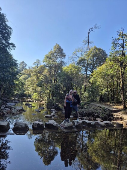 Two people standing on rocks by a calm river surrounded by dense trees. 5