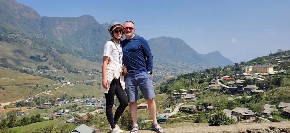 A couple standing on a hill overlooking a village with mountains in the background. 1