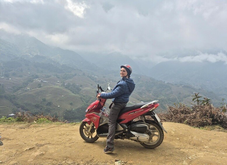 A person sitting on a red motorcycle with a helmet, overlooking a hilly landscape with terraced fields. 3