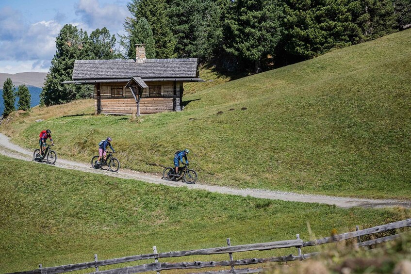 Three cyclists on a dirt path with a wooden cabin in the background. 4