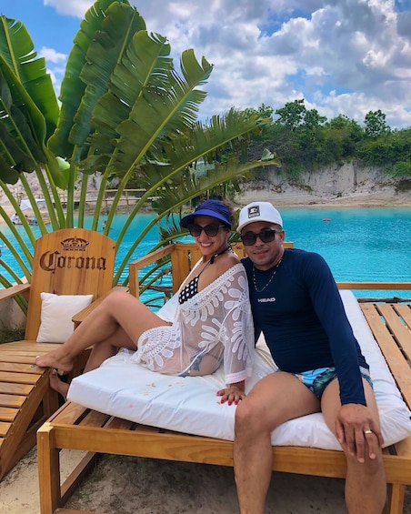 Two people relaxing on a wooden lounge chair by a pool with a wooden sign that reads 'Corona.' 3