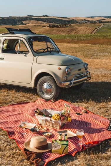 A vintage car parked in a field with a picnic setup on a red and white checkered blanket. 1