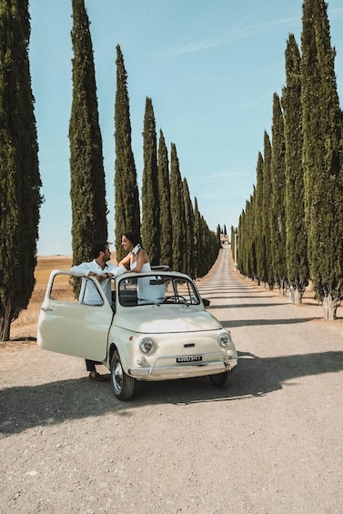 A vintage car parked on a gravel road flanked by tall, narrow trees. 2
