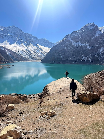 A person walking on a rocky shore with a lake and snow-capped mountains in the background. 1