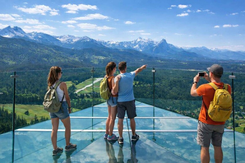 A couple and a photographer on a glass floor overlooking a mountainous landscape. 3