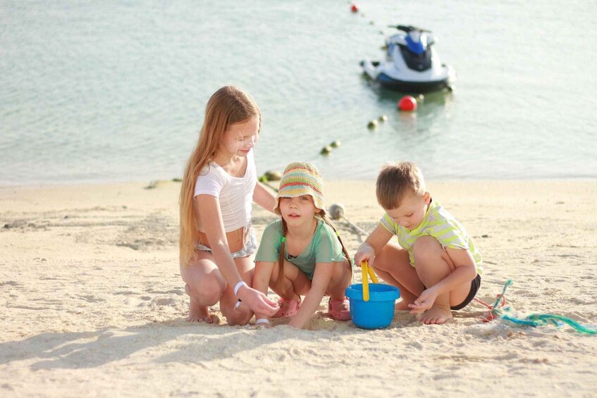 Three children playing with sand and a blue bucket on a beach with a boat in the background. 3
