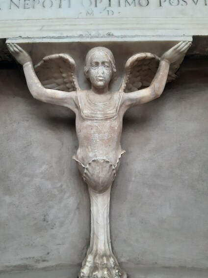 En marmorskulptur af en figur med udstrakte arme, muligvis et religiøst symbol, foran en enkel baggrund. 2