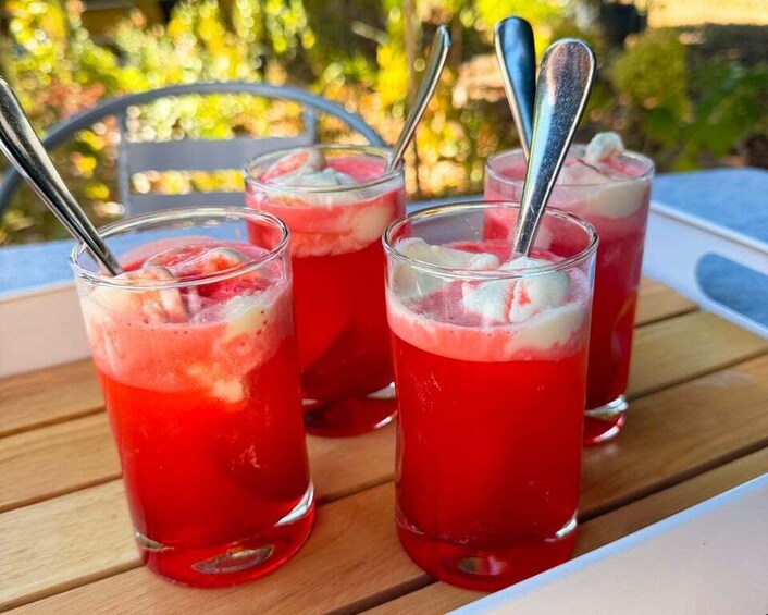 Three glasses of red beverage with ice and a spoon, placed on a wooden table outdoors. 2