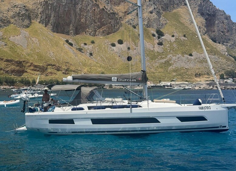 A white sailboat named 'OUFOUR 41' docked near a rocky coastline with a mountainous backdrop. 1