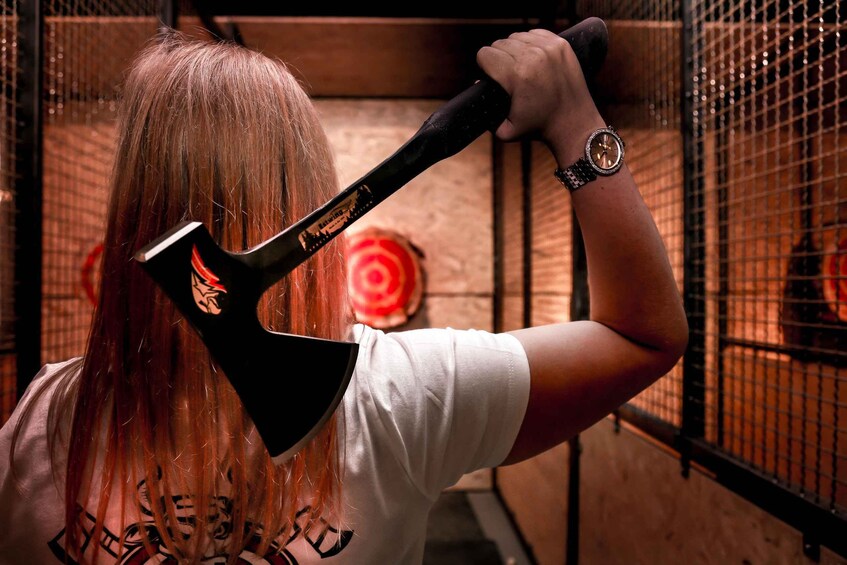 A person holding a black axe with a red and black design, wearing a white t-shirt with black text, in a cage-like structure with a target in the background. 4