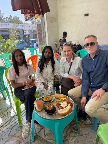 Four people seated at a table with food and drinks, outdoors with plastic chairs and a building in the background. 3