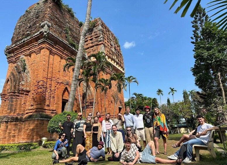 A group of people posing in front of a large, ancient brick structure with two prominent towers, surrounded by palm trees and greenery. 3