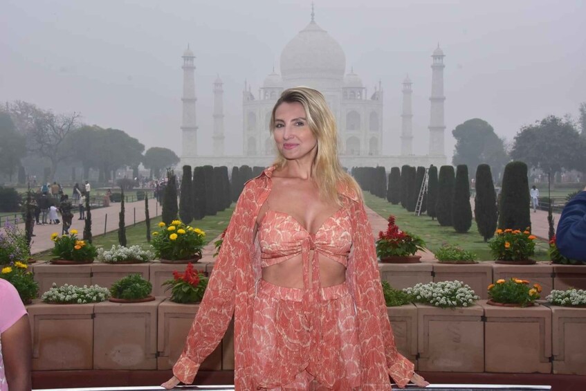 A woman in traditional attire standing in front of a garden with potted plants and a backdrop of a famous white marble mausoleum. 1