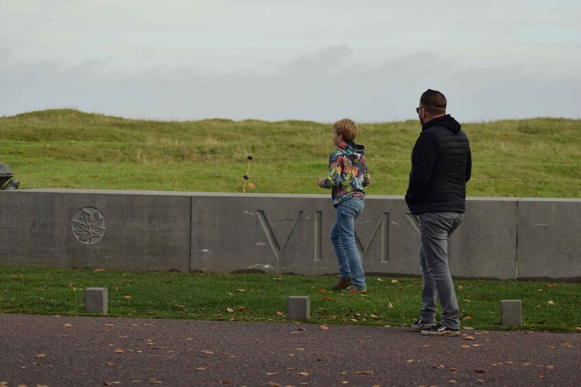 Two people standing by a concrete wall with 'VIM' engraved on it, in a grassy area with fallen leaves on the ground. 3