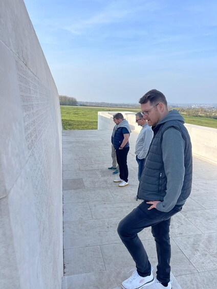A group of people standing on a concrete platform with a large wall featuring inscriptions, overlooking a grassy field and distant buildings. 4