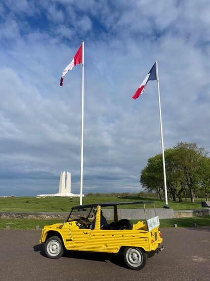 A yellow vintage car parked in a parking lot with two flags on poles in the background. 1