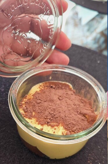 A hand holding a glass container with a powdery substance, possibly cocoa powder, over a bowl with a yellow base, likely a dessert. 4