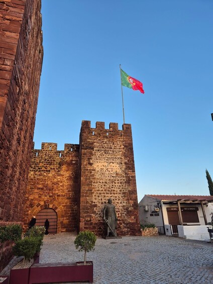 A historic stone tower with a flag, a statue, and cobblestone pavement. 1