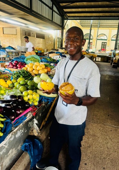 A person holding a yellow fruit at a market stall with various vegetables and fruits displayed. 2