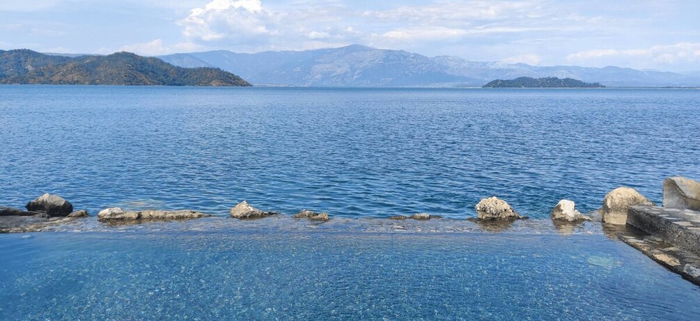 A calm lake with clear blue water, surrounded by rocky shores and distant mountains. 2