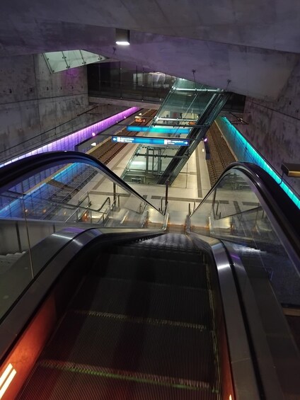An escalator in a subway station with overhead train tracks and signage. 1