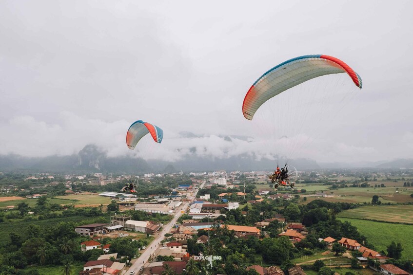 Two paragliders flying over a town with visible buildings and greenery. 2