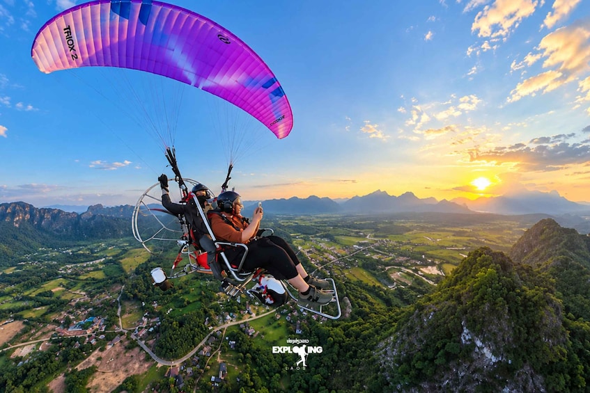 A paraglider with a purple canopy flying over a mountainous landscape during sunset. 1