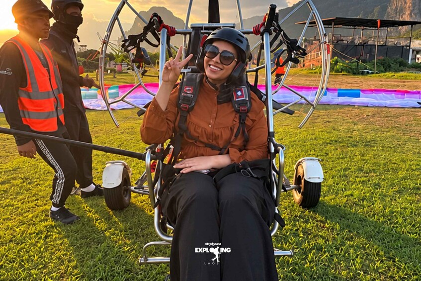 A person in a harnessed chair with a parachute, another person in safety gear, and a grassy field with mountains in the background. 3