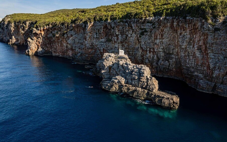 A rugged coastline with a rocky outcrop and a small building on top, surrounded by clear blue waters and green hills. 2